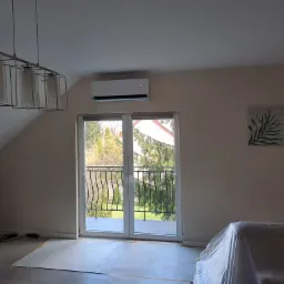 Klimatyzator ścienny zamontowany nad drzwiami balkonowymi w pokoju na poddaszu, meble zabezpieczone folią malarską, widoczna lampa wisząca i obrazek z motywem roślinnym na ścianie.