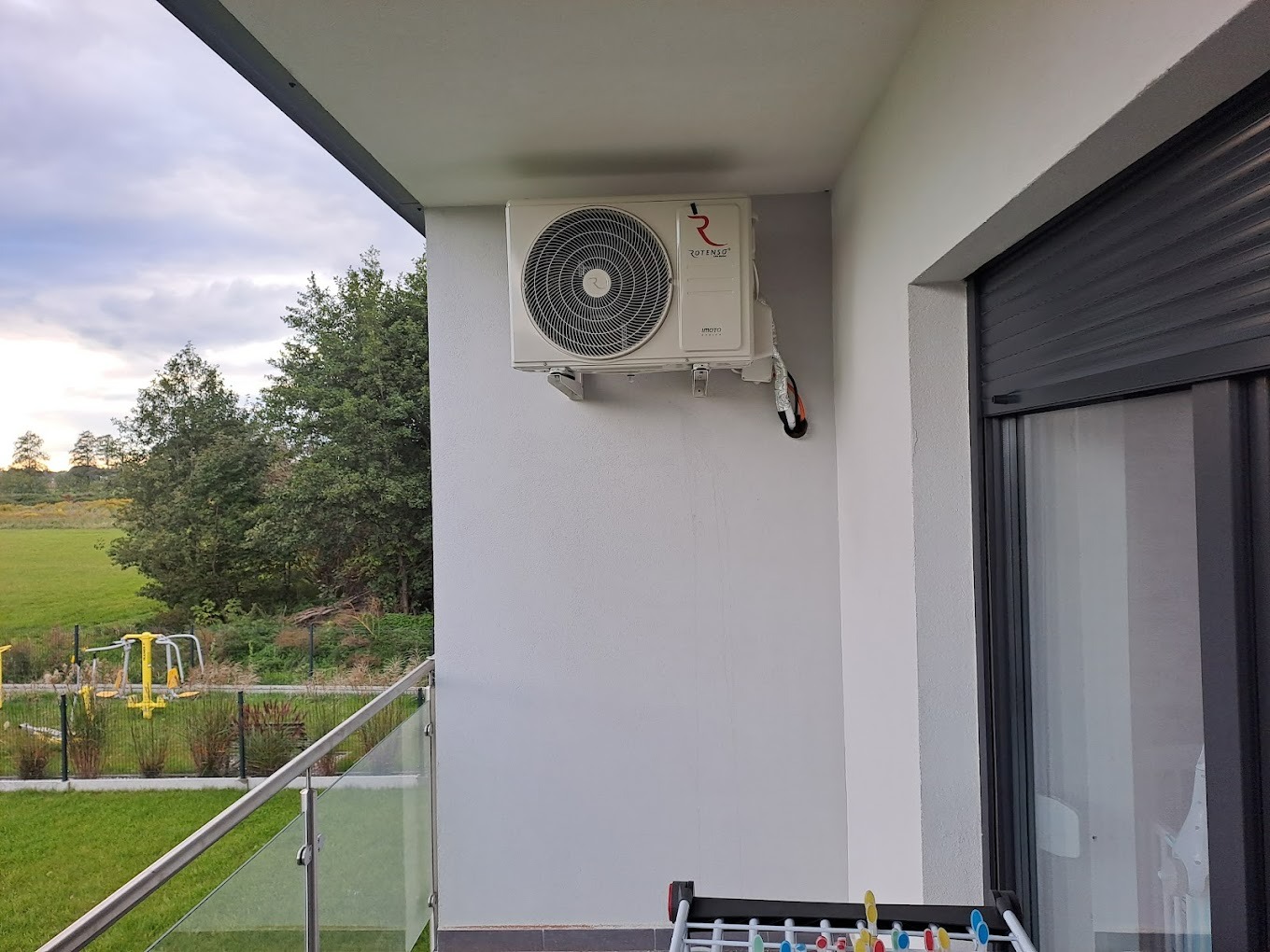 Biała jednostka zewnętrzna klimatyzacji zamontowana na ścianie balkonu z widokiem na zielony krajobraz i plac zabaw