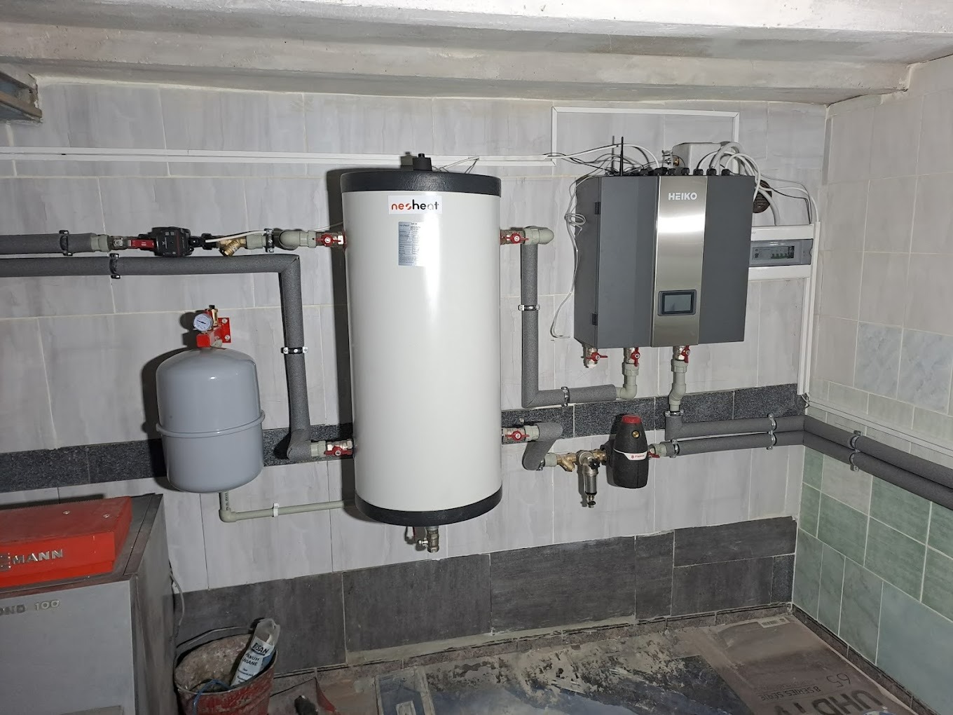 Instalacja grzewcza w kotłowni: biały zasobnik ciepłej wody Nesheat, szara centrala grzewcza Heiko, rury w izolacji, zbiornik wyrównawczy, armatura, stara terakota na ścianach i podłodze.