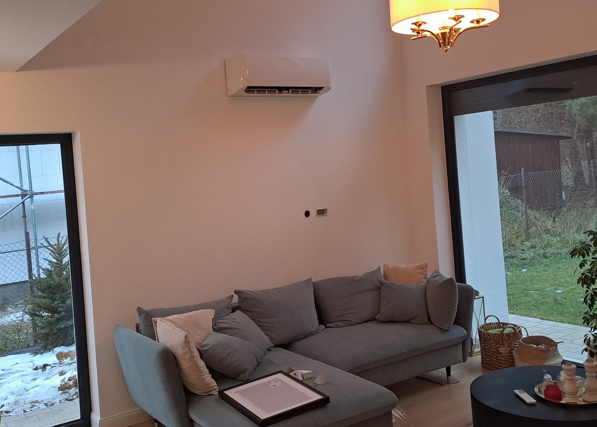 Biały klimatyzator ścienny zamontowany w nowoczesnym salonie z szarą sofą, widok na ogród przez duże okno.