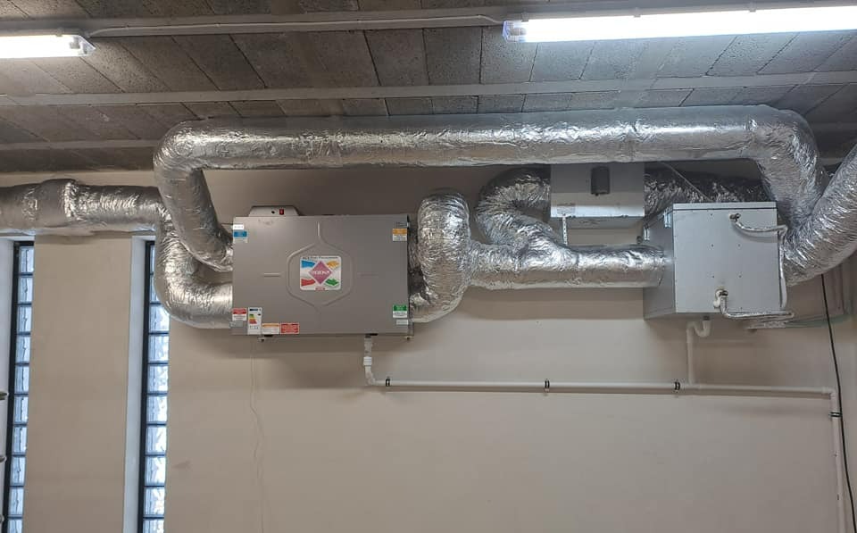 Szara jednostka centralna rekuperacji z aluminiowymi, izolowanymi przewodami wentylacyjnymi, zamontowana pod betonowym stropem w pomieszczeniu z oknem. Widoczne białe rury i przewody elektryczne.