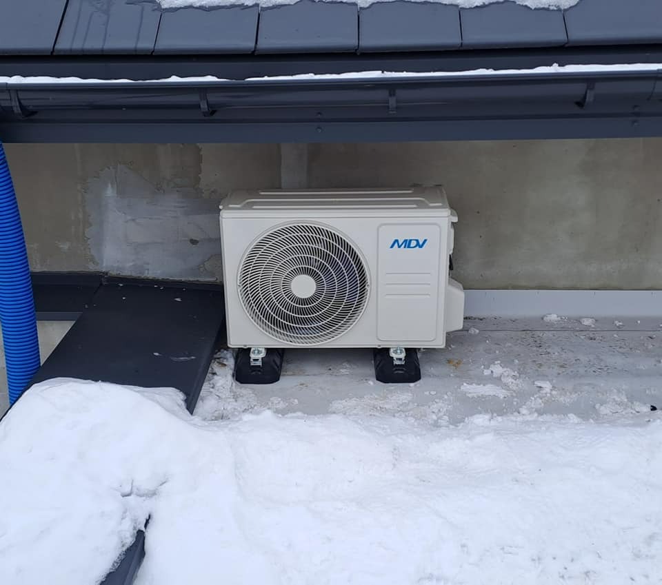 Zewnętrzna jednostka klimatyzacji marki MDV, zamontowana na czarnych podkładkach na ośnieżonym tarasie pod okapem dachu.