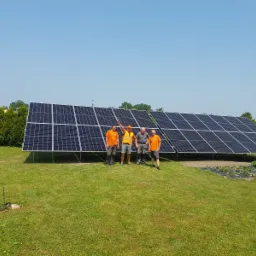 Instalacja fotowoltaiczna na gruncie, ekipa czterech monterów w pomarańczowych koszulkach pozuje na tle paneli, widoczne kable i narzędzia na trawie, w tle zieleń drzew i błękitne niebo.