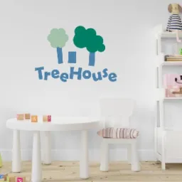 Niebieski napis 'TreeHouse' z dwoma stylizowanymi drzewami w odcieniach zieleni, naklejony na białej ścianie w pokoju dziecięcym, widoczny biały stolik z drewnianymi klockami i krzesło oraz regał...