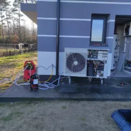 Instalacja zewnętrznej jednostki urządzenia grzewczego przy szarym budynku z widocznym wnętrzem. W tle narzędzia, skrzynka narzędziowa i ciekawski kot.