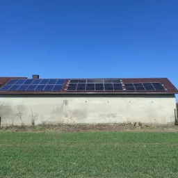 Panele słoneczne zamontowane na dachu budynku gospodarczego z szarą elewacją, widok z poziomu trawnika w słoneczny dzień.