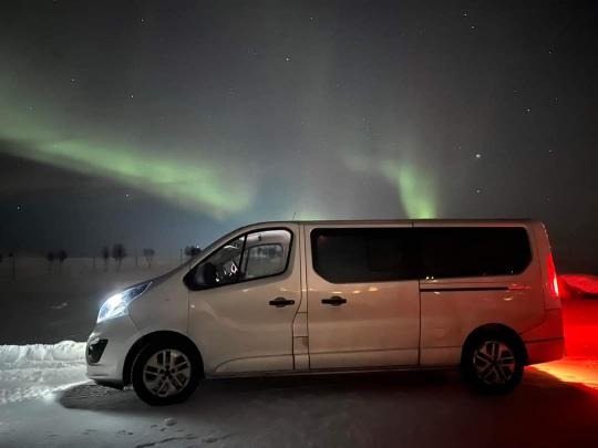 Biały bus na tle zorzy polarnej i nocnego nieba usianego gwiazdami, zaparkowany na ośnieżonym terenie.