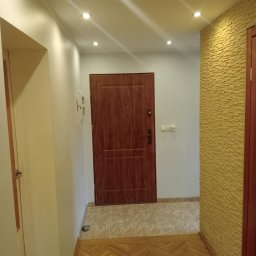 KOBRAStyle Maciej Wąsikowski - Korytarz po remoncie: drewniana podłoga w jodełkę, brązowe drzwi wejściowe, ściana z dekoracyjną okładziną w odcieniach żółci, punktowe oświetlenie sufitowe.