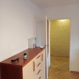 KOBRAStyle Maciej Wąsikowski - Jasne wnętrze mieszkania po malowaniu: pomalowane na biało ściany, parkiet w jodełkę, komoda z brązowym blatem i otwarta biała rama drzwi z widokiem na ścianę z dekoracyjną okładziną.