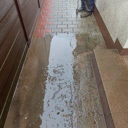 KOBRAStyle Maciej Wąsikowski - Mężczyzna w roboczych butach czyści kostkę brukową za pomocą myjki ciśnieniowej, woda zbiera się na powierzchni, tworząc kałuże z resztkami brudu i liści.