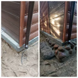 Fragment drewnianej konstrukcji z brązową rynną, osadzonej na betonowej podstawie, obok której widoczna jest ziemia z kamieniami i piasek.