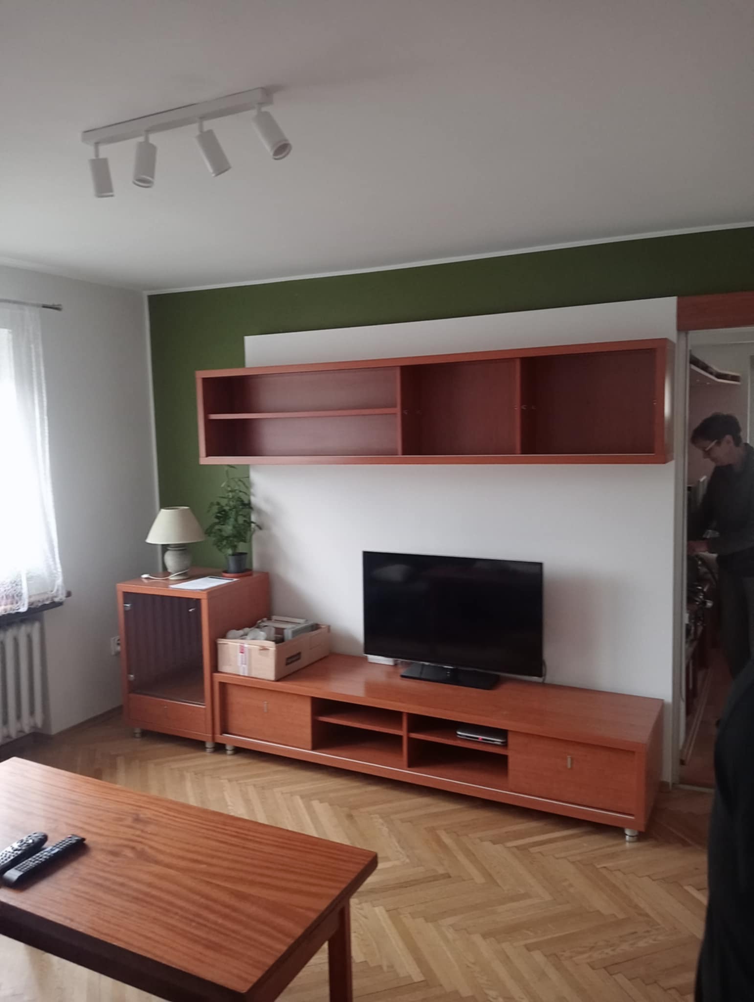 Pokój dzienny z drewnianą meblościanką, telewizorem, stolikiem kawowym i parkietem ułożonym w jodełkę; ściana pomalowana częściowo na zielono.