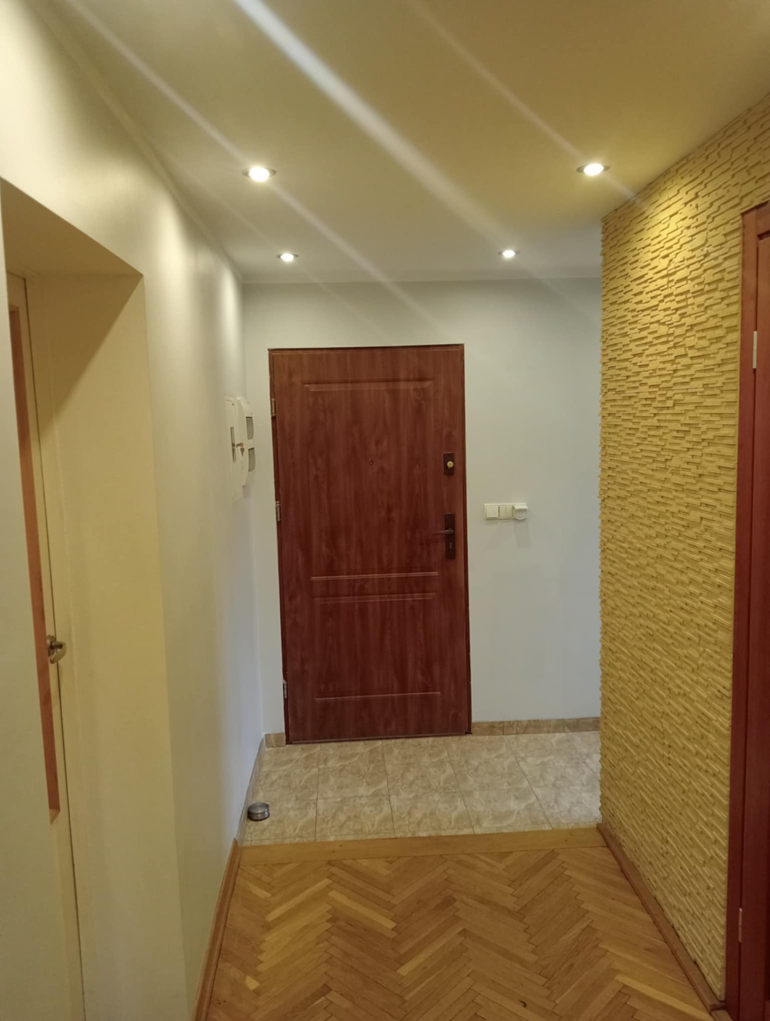 Korytarz po remoncie: drewniana podłoga w jodełkę, brązowe drzwi wejściowe, ściana z dekoracyjną okładziną w odcieniach żółci, punktowe oświetlenie sufitowe.