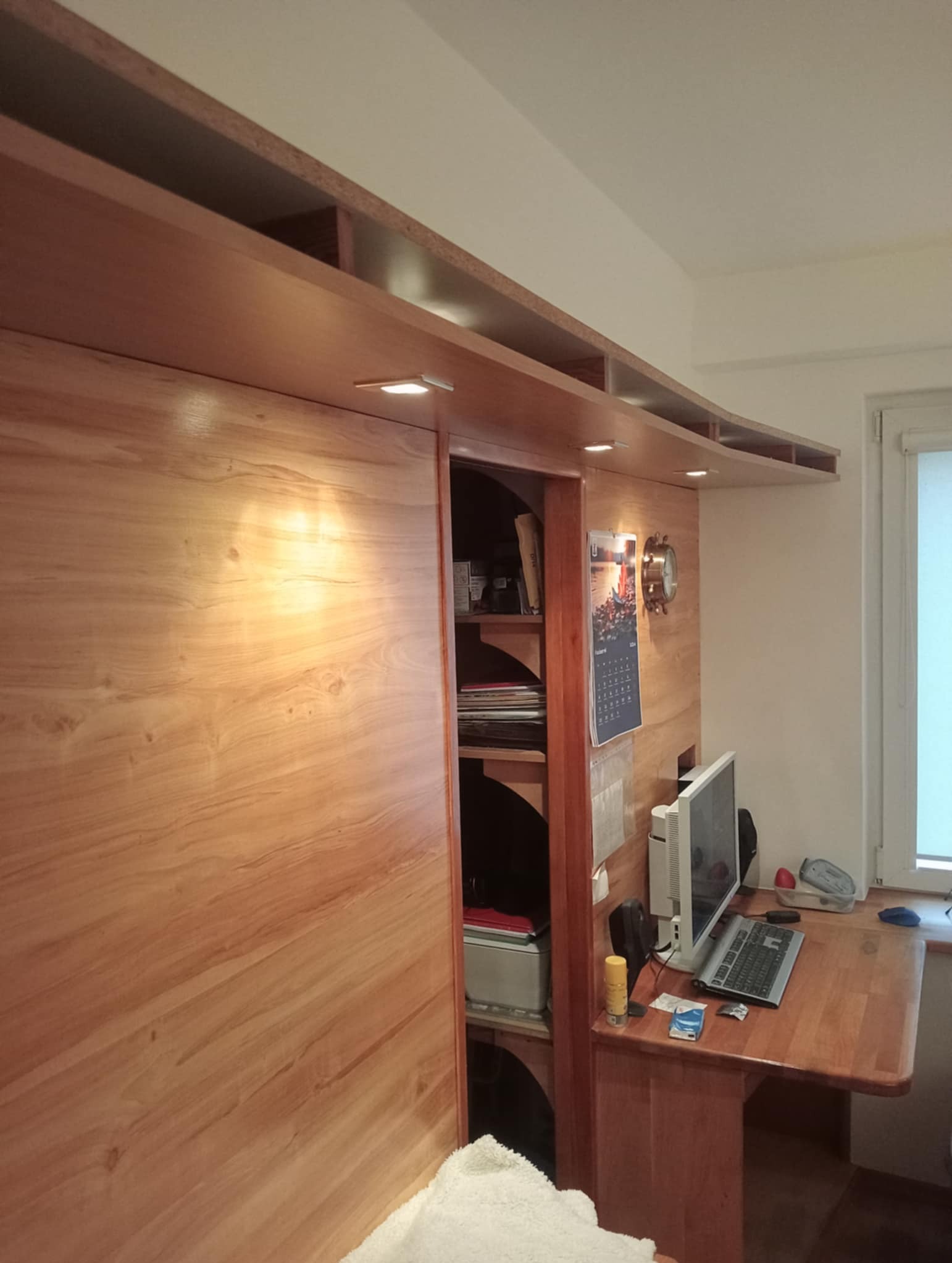 Drewniana zabudowa meblowa ze schowkami i oświetleniem punktowym oraz biurkiem w Stargardzie. Widoczne półki z książkami i kalendarz z zegarem.