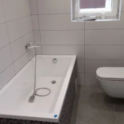 Nowoczesna łazienka z białą wanną obudowaną mozaiką, jasnymi płytkami ściennymi, szarą podłogą i podwieszaną toaletą, oświetlona punktowo.