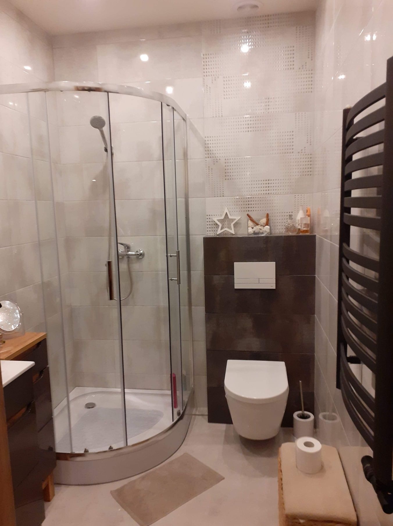 Nowoczesna łazienka z kabiną prysznicową narożną, wiszącą toaletą na tle brązowych płytek i grzejnikiem drabinkowym w ciemnym kolorze.