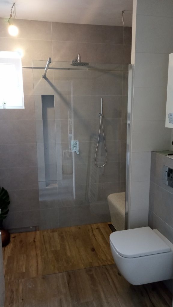 Nowoczesna łazienka z kabiną prysznicową ze szklaną ścianką, drewnopodobną podłogą i wiszącą toaletą.