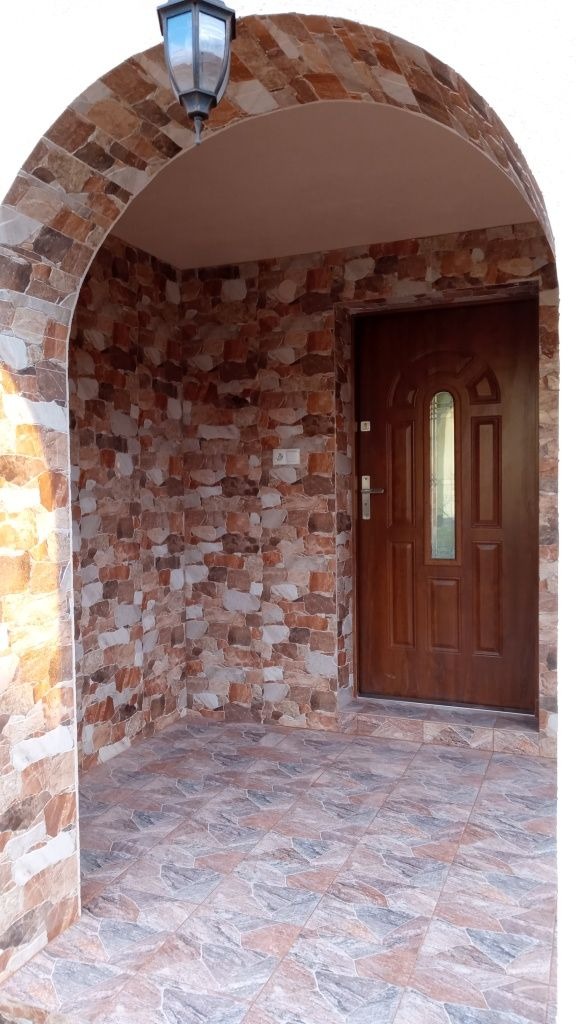 Zadaszone wejście do domu z brązowymi płytkami imitującymi kamień na ścianach i podłodze, oświetlone lampą sufitową nad drewnianymi drzwiami z szybą.