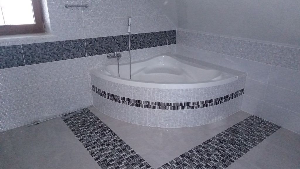 Narożna wanna w łazience wykończonej jasnymi płytkami z dekoracyjnymi pasami mozaiki w odcieniach szarości i czerni na ścianach i podłodze.