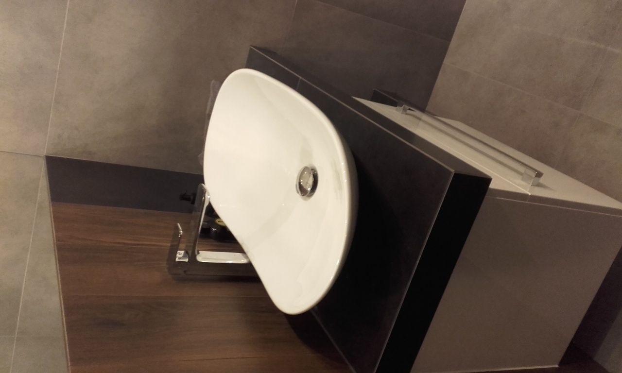 Nowoczesna, biała umywalka nablatowa o asymetrycznym kształcie, zamontowana na ciemnobrązowej szafce w łazience z szarymi płytkami i drewnianą podłogą.