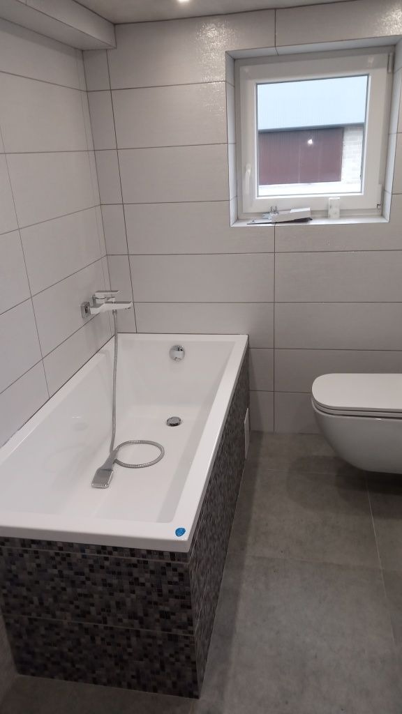 Nowoczesna łazienka z białą wanną obudowaną mozaiką, jasnymi płytkami ściennymi, szarą podłogą i podwieszaną toaletą, oświetlona punktowo.