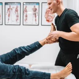 Mężczyzna w czarnym ubraniu wykonuje manualną terapię stopy pacjenta leżącego na białej leżance w gabinecie, na ścianie wiszą plakaty anatomiczne.
