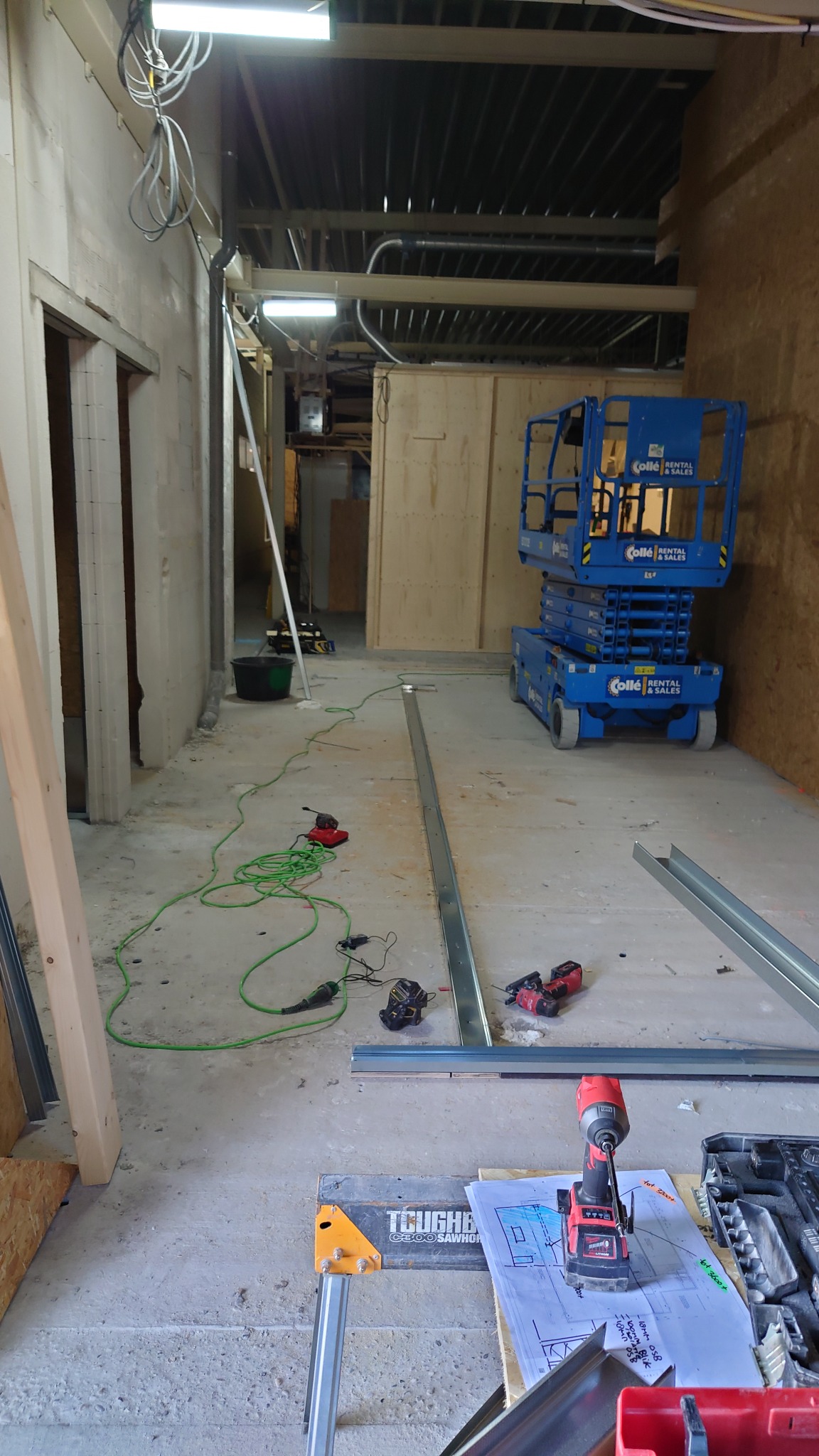 Wnętrze w trakcie remontu z widocznymi profilami metalowymi, podnośnikiem nożycowym, narzędziami akumulatorowymi, przewodami elektrycznymi i planami na stole warsztatowym.