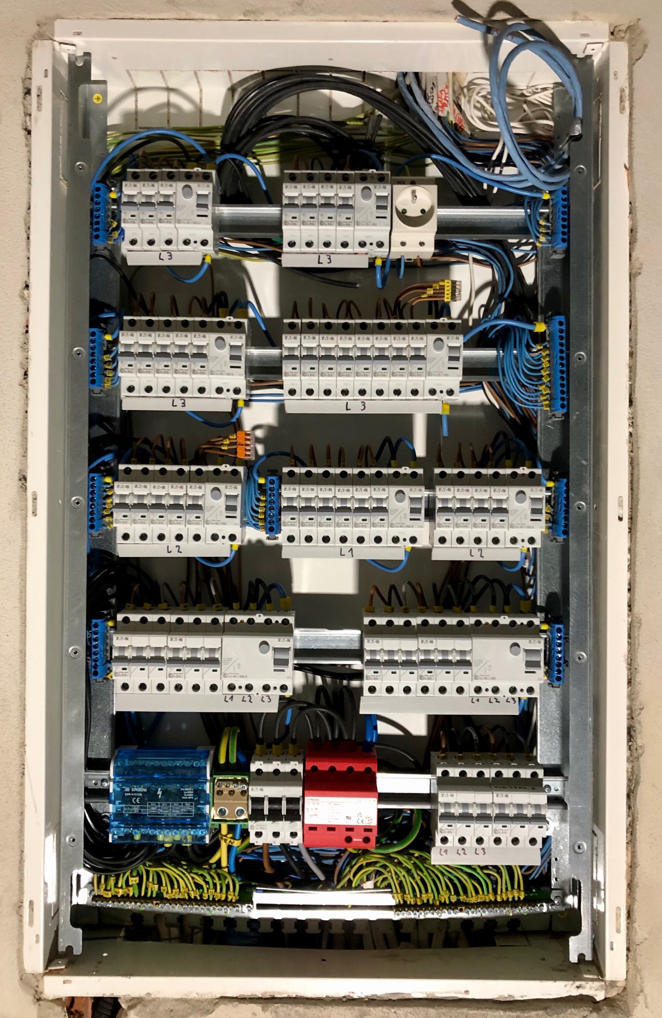 Wnętrze rozdzielnicy elektrycznej z zamontowanymi zabezpieczeniami przepięciowymi Dehn (typ 1 i 2) w kolorze czerwonym, wyłącznikami różnicowoprądowymi Eaton oraz modułem Ergom w kolorze niebieskim...