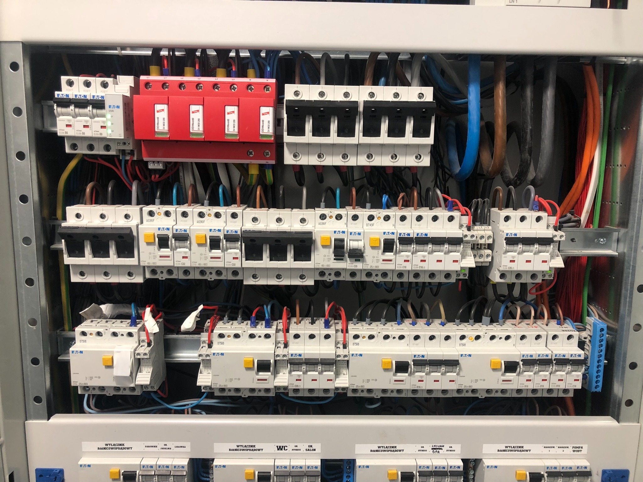 Wnętrze rozdzielnicy elektrycznej z widocznymi zabezpieczeniami przeciwprzepięciowymi (czerwone moduły Dehnventil), wyłącznikami różnicowoprądowymi Eaton oraz licznymi przewodami o różnych kolorach.
