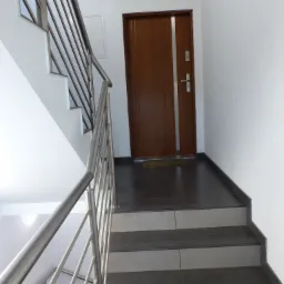 Klatka schodowa w nowoczesnym budynku z metalową balustradą i brązowymi drzwiami wejściowymi.