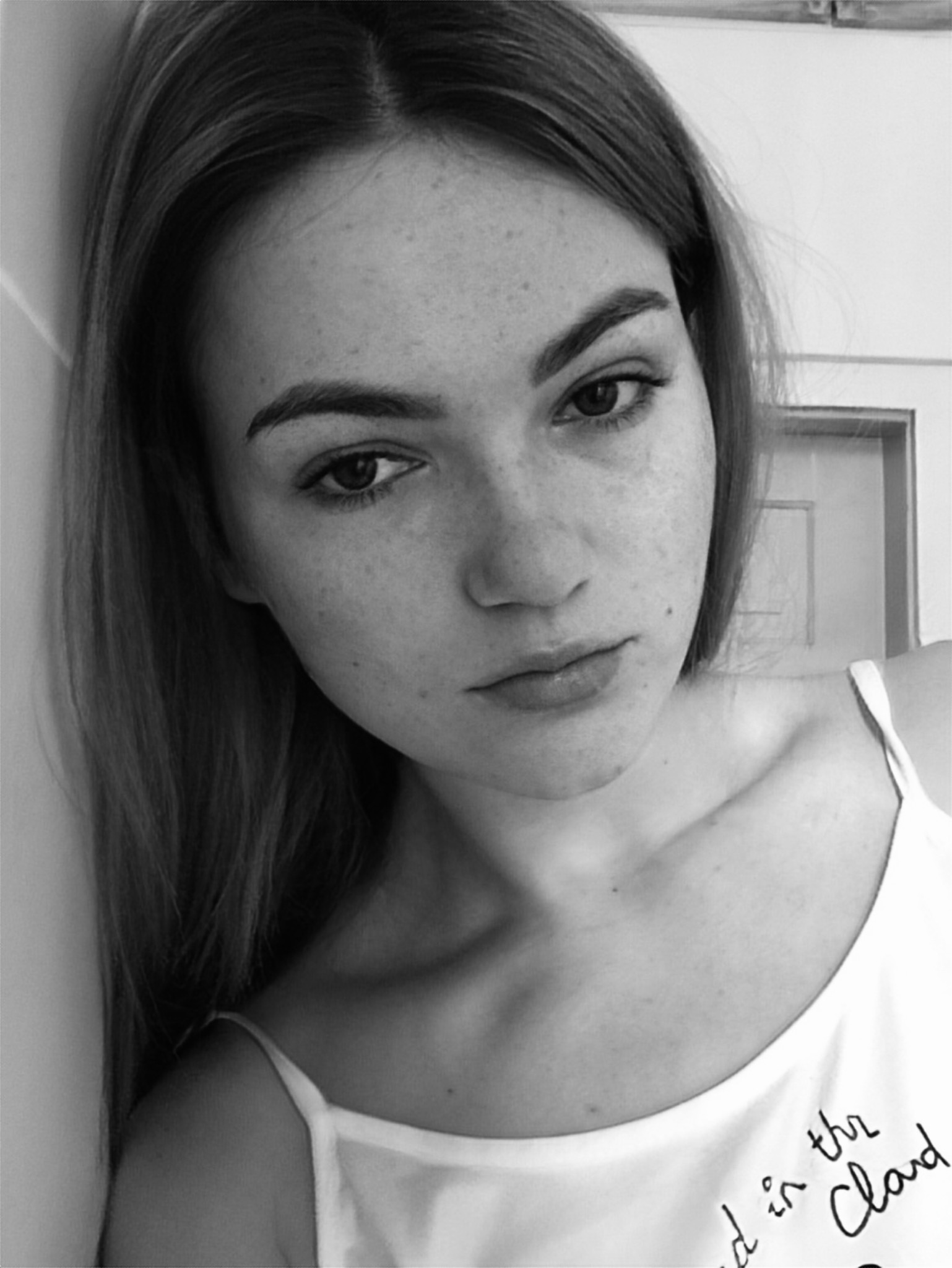 Portret młodej kobiety z piegami, oparta o ścianę, w czarno-białej tonacji, z napisem na koszulce.