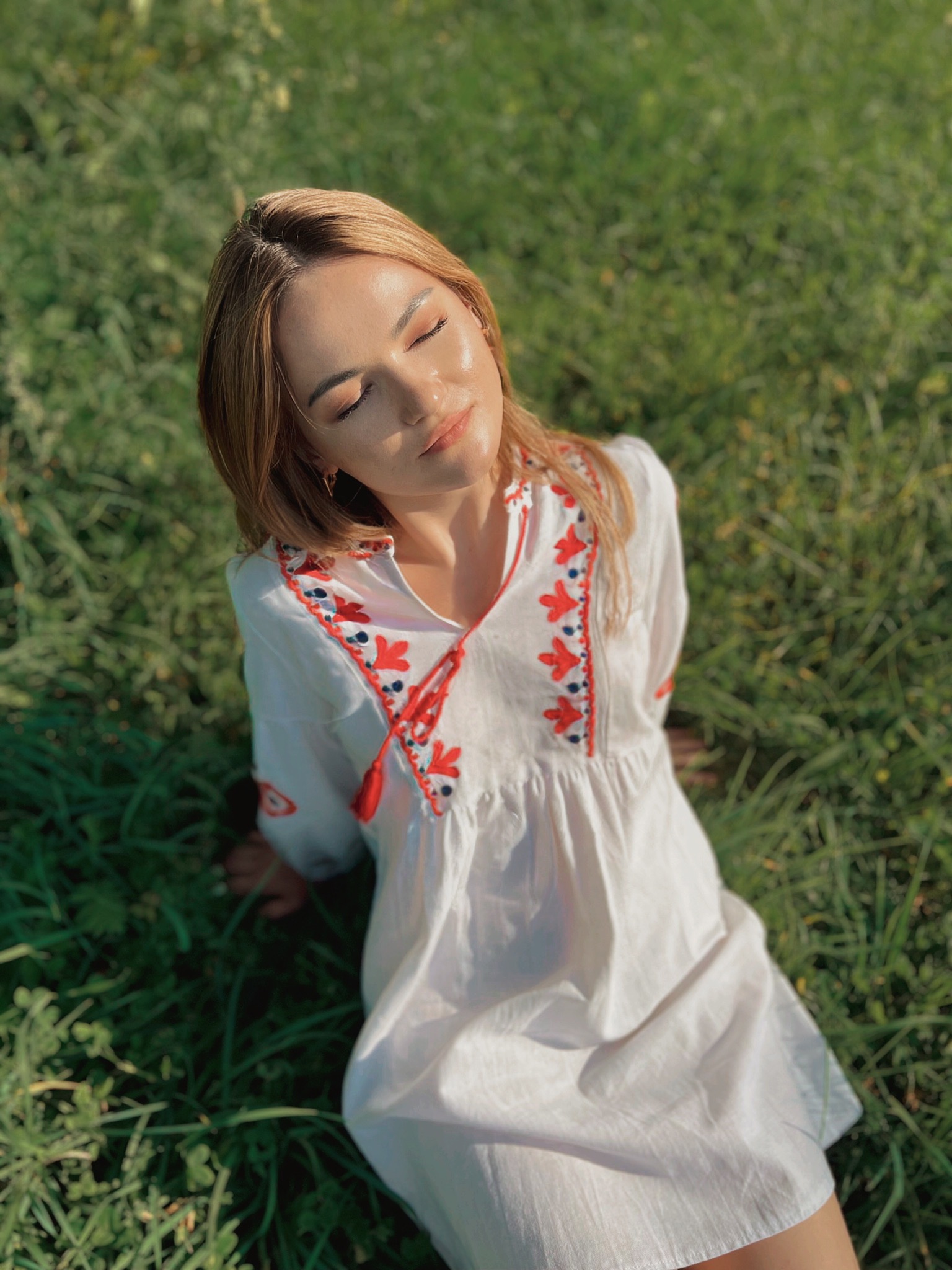 Młoda kobieta w białej sukience z haftem, siedząca na trawie z zamkniętymi oczami, ogrzewana promieniami słońca.