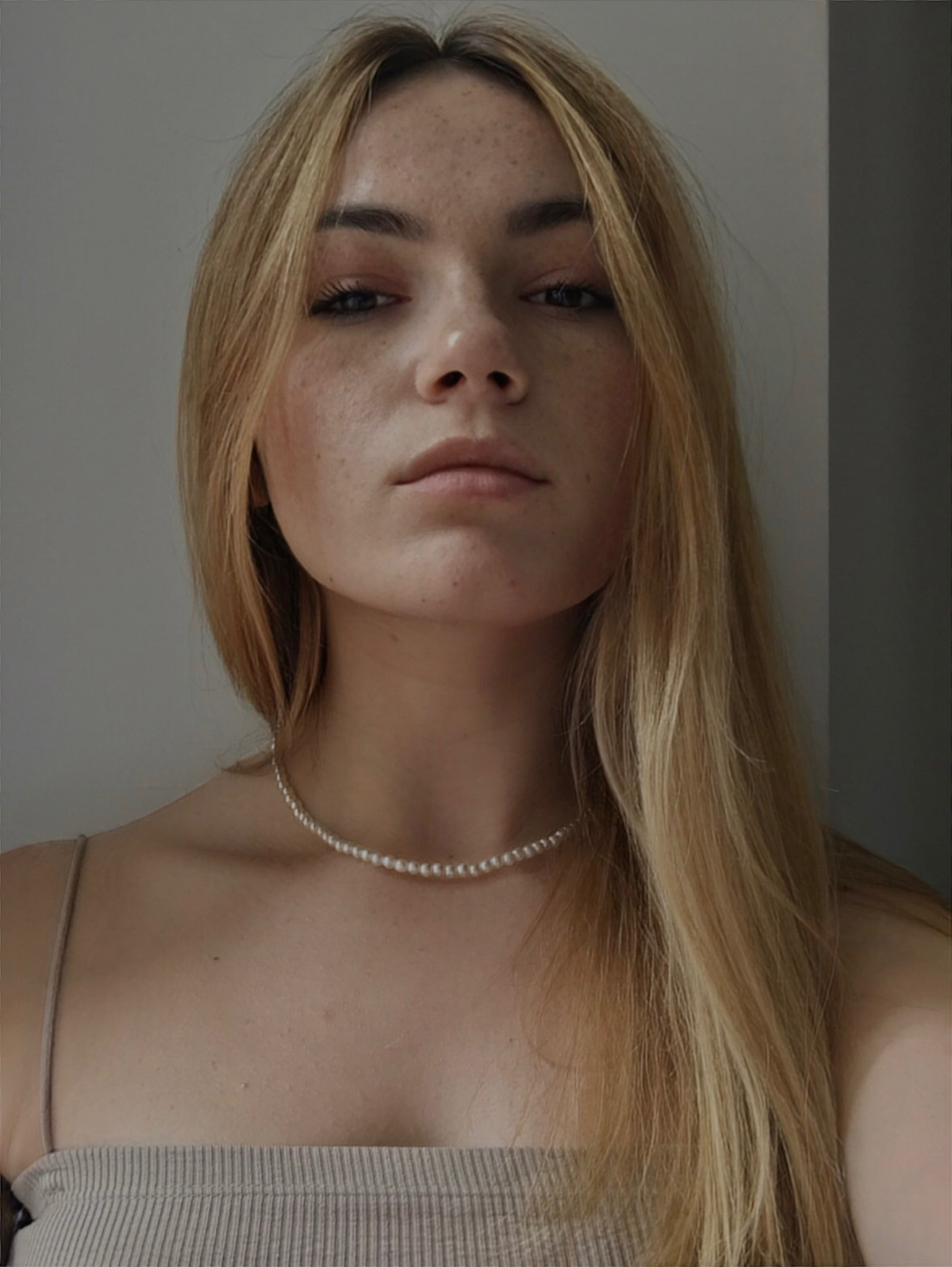 Portret młodej kobiety z blond włosami, delikatnym makijażem i perłowym naszyjnikiem, patrzącej prosto w obiektyw w neutralnym otoczeniu.