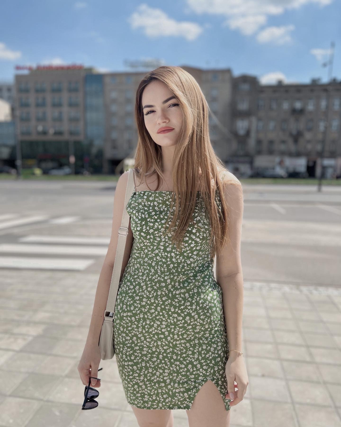 Kobieta w zielonej sukience w białe kwiatki, stojąca na tle miejskiego krajobrazu z budynkami i przejściem dla pieszych.