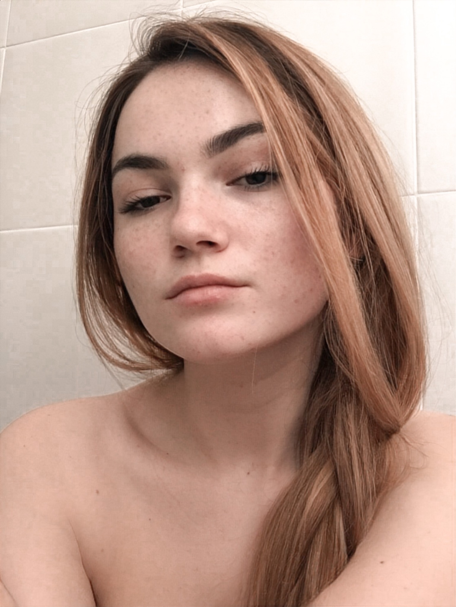 Portret młodej kobiety z piegami, o długich, zaplecionych w warkocz włosach w odcieniach blondu i brązu, na tle białych płytek.