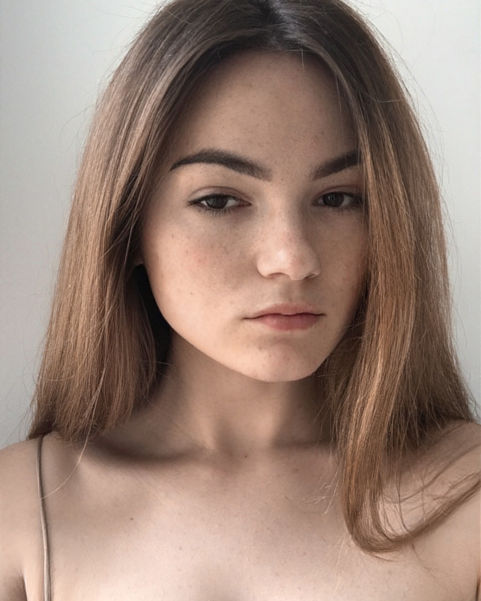 Portret młodej kobiety z długimi, brązowymi włosami, delikatnym makijażem i piegami na twarzy, sfotografowanej na jasnym tle.