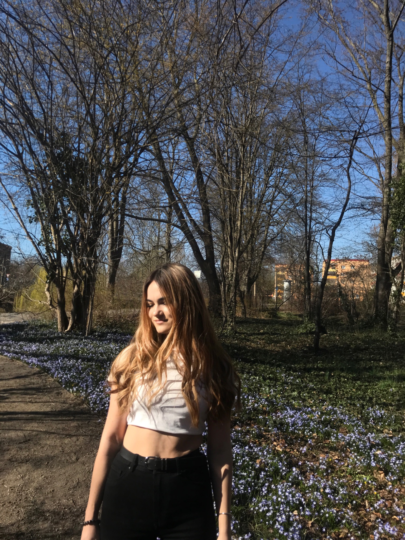 Młoda kobieta w białym topie i czarnych spodniach pozuje na tle kwitnących niebieskich kwiatów i drzew bez liści w słoneczny dzień.