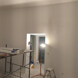 BEC-BUD FIRMA USŁUGOWO-HANDLOWA KAROL ZAGDAN - Ściana w trakcie szpachlowania z widocznym rusztowaniem, lampą halogenową i narzędziami. Fragment dłoni w lewym dolnym rogu kadru. W tle widoczne drzwi.