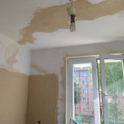 BEC-BUD FIRMA USŁUGOWO-HANDLOWA KAROL ZAGDAN - Pokój w trakcie szpachlowania: fragmenty ścian i sufitu pokryte szpachlą, widoczne okno z widokiem na blok. Żarówka zwisa z sufitu.