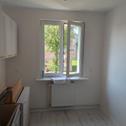 BEC-BUD FIRMA USŁUGOWO-HANDLOWA KAROL ZAGDAN - Jasne wnętrze po malowaniu: białe ściany, okno z widokiem na zieleń, grzejnik pod oknem, częściowo widoczne białe szafki kuchenne z złotymi uchwytami.