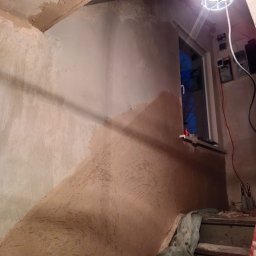 BEC-BUD FIRMA USŁUGOWO-HANDLOWA KAROL ZAGDAN - Ściana w trakcie szpachlowania, widoczne nierówności i warstwy gładzi. Na górze lampa budowlana, a w tle okno. Fragment schodów z folią malarską.