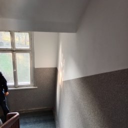BEC-BUD FIRMA USŁUGOWO-HANDLOWA KAROL ZAGDAN - Klatka schodowa po malowaniu: białe ściany u góry, szare u dołu. Widoczne schody i okno z widokiem na drzewa. Promienie słońca na ścianie.