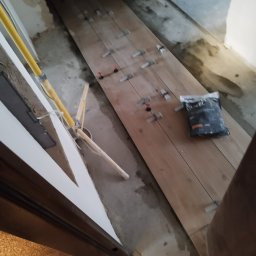 BEC-BUD FIRMA USŁUGOWO-HANDLOWA KAROL ZAGDAN - Układanie płytek imitujących drewno w wąskim korytarzu, z użyciem systemu poziomowania. Widoczny worek z klejem i lampa robocza.