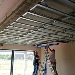 BEC-BUD FIRMA USŁUGOWO-HANDLOWA KAROL ZAGDAN - Montaż sufitu podwieszanego z płyt karton-gips: ekipa na drabinach mocuje konstrukcję z profili, widoczne płyty w kolorze różowym, duże okno w tle.