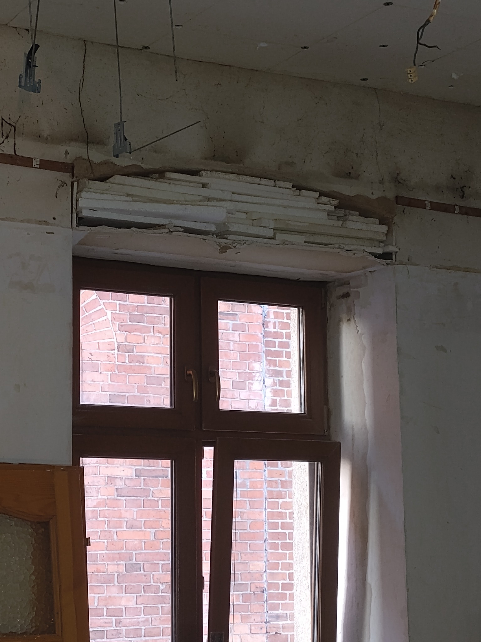 Fragment remontowanego wnętrza: widoczne płyty izolacyjne nad oknem, surowy tynk i instalacje sufitowe w trakcie prac wykończeniowych. Ceglany budynek za oknem.