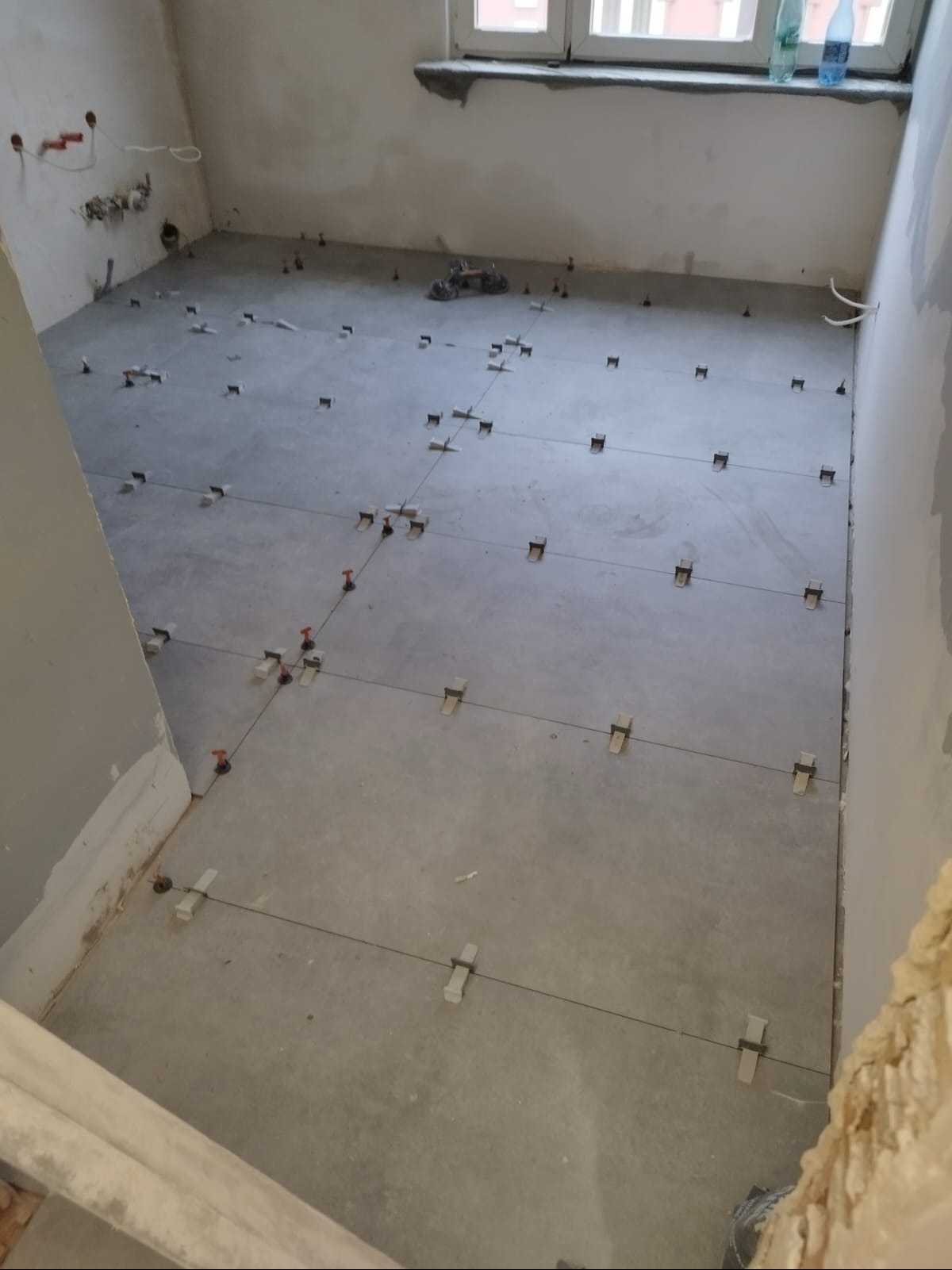 Przygotowanie do układania płytek podłogowych w jasnym pomieszczeniu. Widoczne poziomowanie z użyciem krzyżyków i sznurka. W tle okno i ściany w stanie surowym.