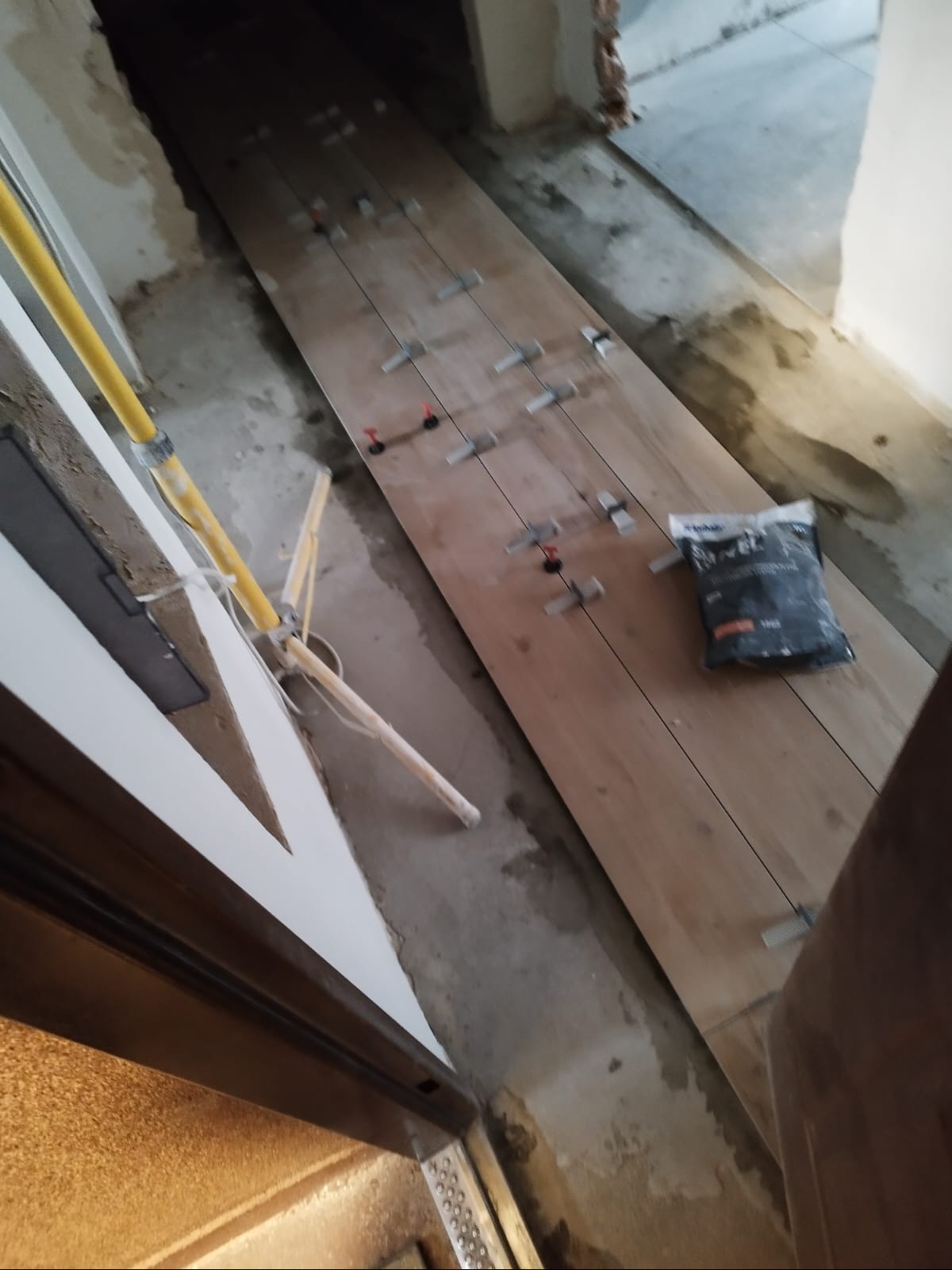 Układanie płytek imitujących drewno w wąskim korytarzu, z użyciem systemu poziomowania. Widoczny worek z klejem i lampa robocza.