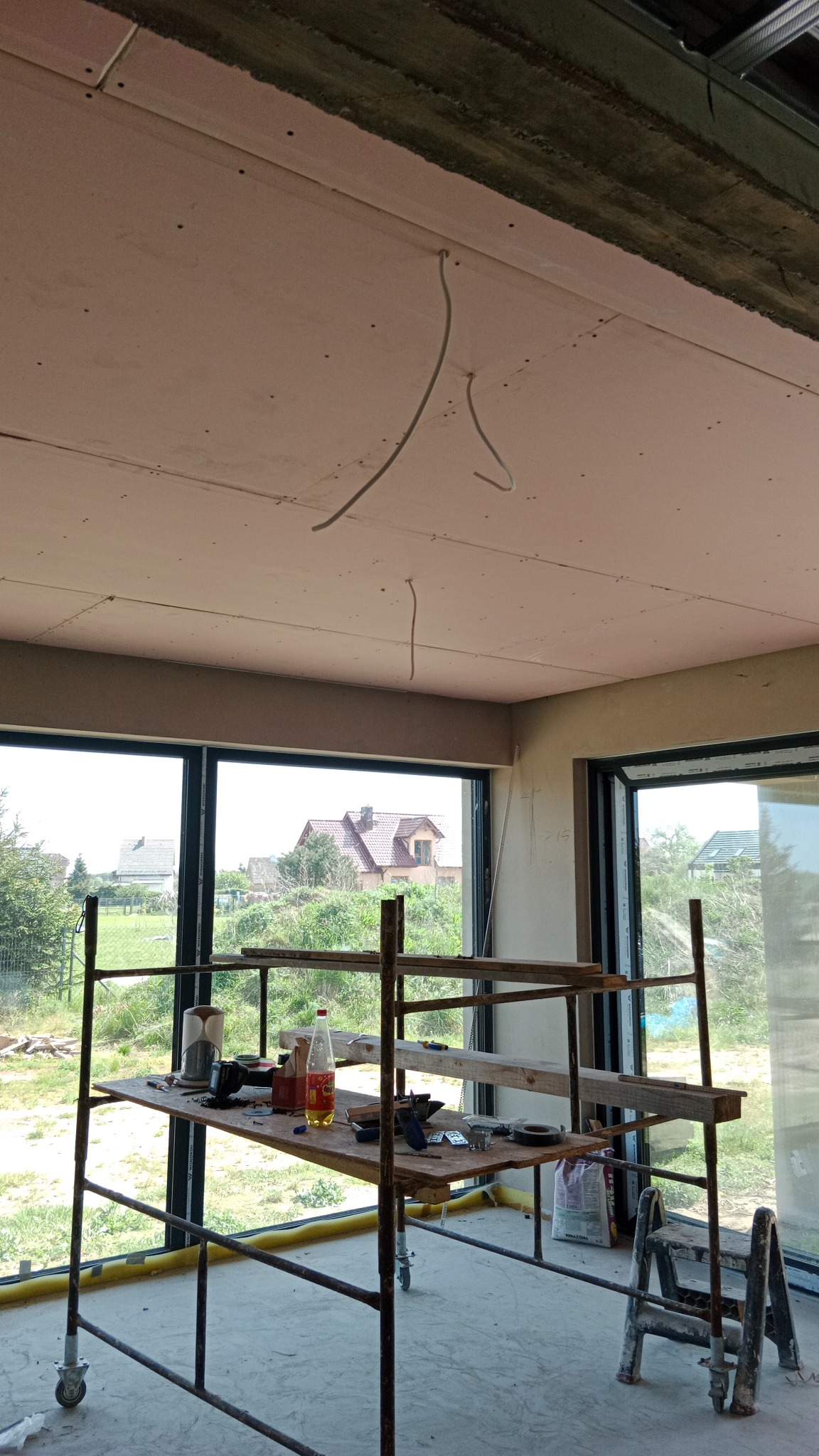 Montaż płyt karton-gips na suficie w nowym domu. Widoczne kable, rusztowanie i narzędzia. Przez okna widać zieleń i domy.