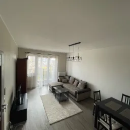 Jasne wnętrze po remoncie: salon z beżową sofą narożną, ciemnym stolikiem kawowym na tle białego dywanu, drewniana podłoga, stół z krzesłami i widok na miasto przez okno.