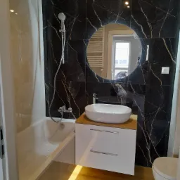 Nowoczesna łazienka z marmurową ścianą, okrągłym lustrem i białą umywalką nablatową z szafką, widoczna wanna i toaleta.